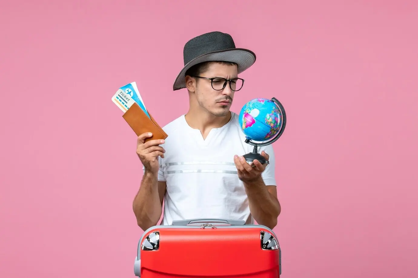 Frontalansicht eines jungen Mannes, der Urlaubstickets und einen kleinen Globus vor pinker Wand hält.