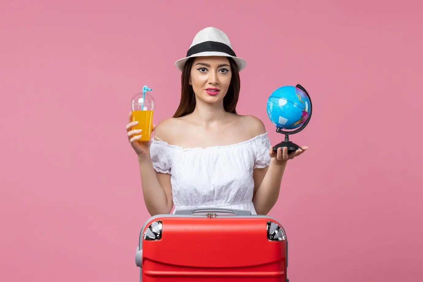 Vorderansicht einer jungen Frau, die einen kleinen Globus und einen Cocktail hält, vor pinkfarbener Wand; Sommer, Reise, Trip, Hitze, Farbe, Urlaub.